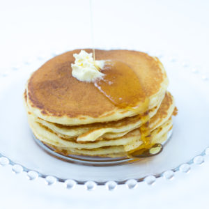 Short stack of cornmeal pancakes made with cornmeal pancake mix with melting butter and syrup being poured onto stack, sitting on glass plate.