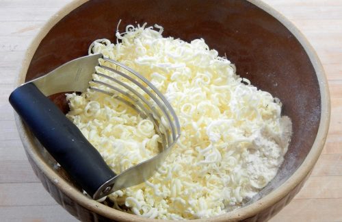 Looking inside a brown mixing bowl; flour mixture on bottom of bowl, and frozen grated butter on top with pastry cutter.