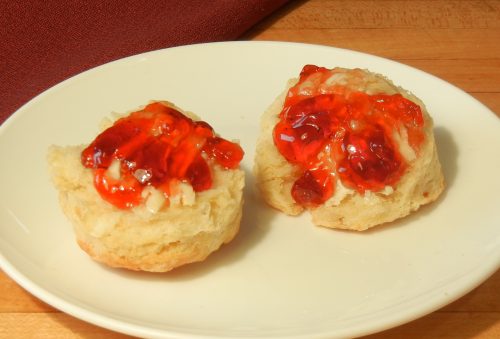 buttermilk biscuit on white china plate, split in half, spread with butter and topped with red jelly.